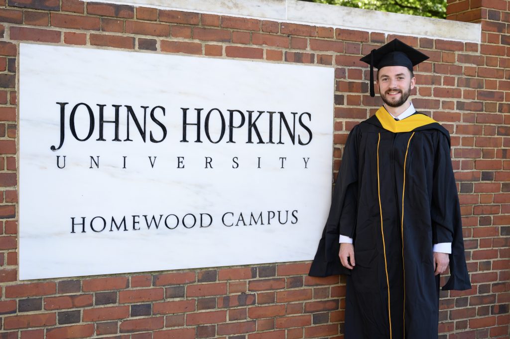 Ryan Demo stands next to the Johns Hopkins University Homewood campus sign in graduation garb.