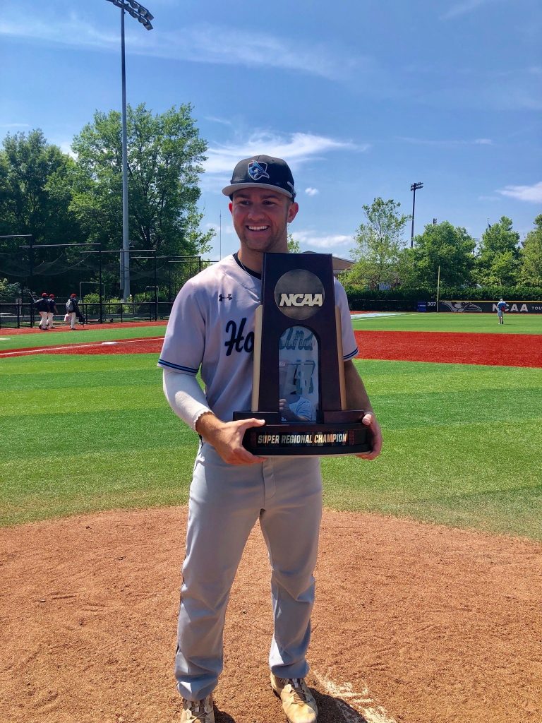 Dressed in a Hopkins baseball uniform and standing on the pitcher's mound of the JHU baseball field, Tim Kutcher holds an NCAA Super Regional Champion award.