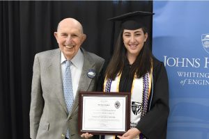 Seymour and Jennifer Baron hold a plaque. Jennifer wears graduation regalia.