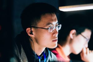 Chenxi Liu in a dark room, illuminated by the light of a computer screen and wearing earphones.