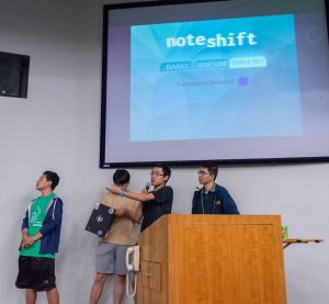 The team members of Noteshift present their work in Hodson 110. A screen displays the Noteshift user interface, labeled "Noteshift: Band, Semitone, Simple Key. Transpose to the key of:" with a purple square.