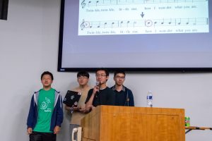 The team members of Noteshift present their work in Hodson 110. Music notes and the lyrics of "Twinkle Twinkle Little Star" are displayed on the projector screen behind them.