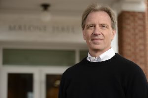 Headshot of Gregory Hager wearing a black sweater over a white shirt standing in front of the front door of Malone Hall.