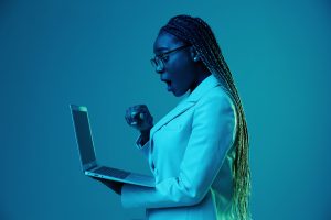 An African American woman wearing glasses and a blazer gasps at a laptop.