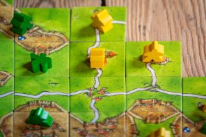 A tile-based board game showing grass and settlements with yellow and green person tokens placed on some of the squares.