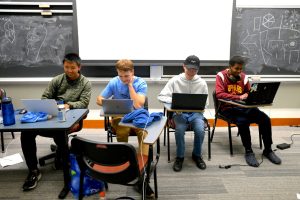 Four students type on laptops, sitting in desks in front of a blackboard.