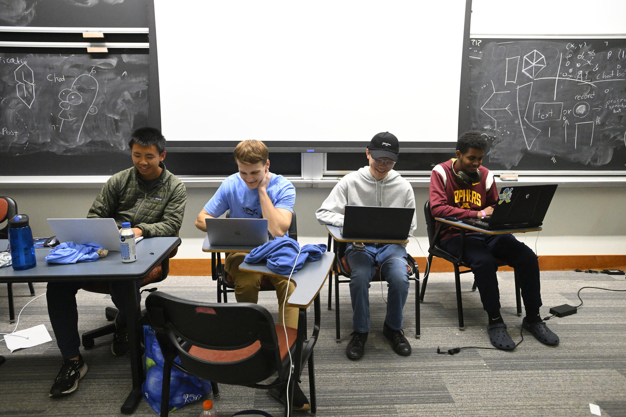 Four students type on laptops, sitting in desks in front of a blackboard.