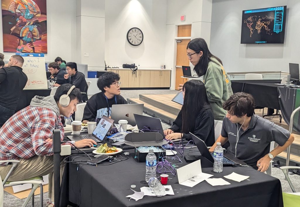 The team huddles around laptops at a table littered with water bottles, cords, and snacks. On the wall there is an analogue clock and a digital map of the world.