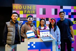 The ChainSplash team at the ETHGlobal Hackathon in front of a backdrop that reads "Nouns fair." One team member wears oversized square cardboard glasses and displays an iPad. Another team member holds up a laptop screen.