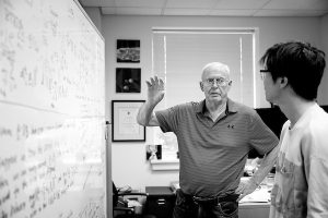 Black-and-white image of Alan Yuille in present day as a professor, shown in front of a white board discussing its contents with a student.