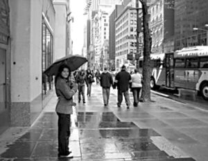 Black-and-white image of a woman holding an umbrella on a rainy busy city street with passersby in the background.